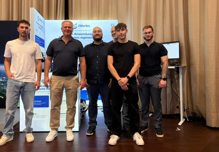 Das iWerbo-Team: sechs Personen stehen nebeneinander vor dem eigenen Messestand mit dem iWerbo-Logo